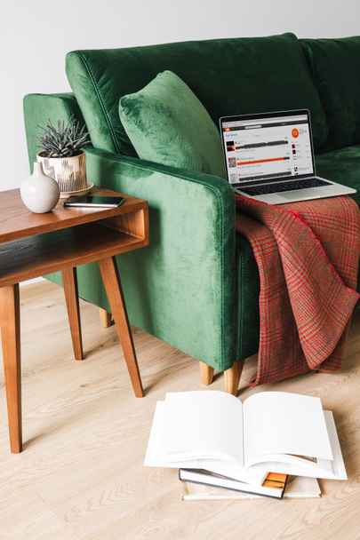 KYIV, UKRAINE - APRIL 14, 2020: green sofa with blanket and laptop with soundcloud website near wooden coffee table with plant and smartphone near books on floor - Foto, Bild