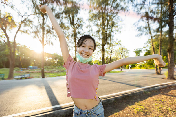 Щаслива усміхнена азіатська дівчинка в масці під підборіддям, що розширюється в зеленій природі, жінка насолоджується диханням свіжого повітря з піднятими руками після карантину Covid-19, Coronavirus закінчився
 - Фото, зображення