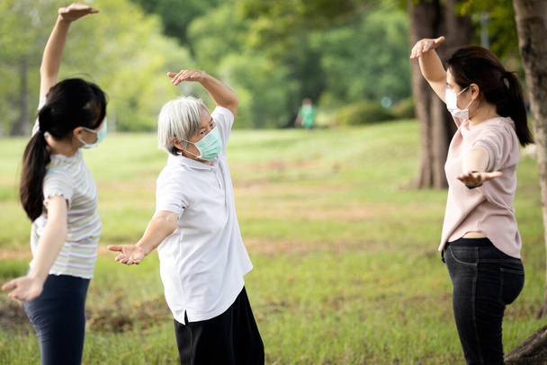 La familia feliz sonriendo mientras hace ejercicio en el parque al aire libre, la madre, la hija y la abuela sana están estirando el ejercicio del brazo, usando máscara quirúrgica, disfrutan, se relajan después de la cuarentena del Coronavirus o Covid-19
 - Foto, Imagen