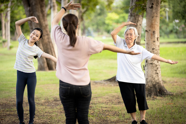 Familia feliz sonriendo mientras hacen ejercicio juntos en el parque al aire libre, madre, hija y abuela sana están estirando el ejercicio del brazo, disfrutar, relajación después de la cuarentena del Coronavirus o Covid-19, cuidado de la salud
 - Foto, Imagen