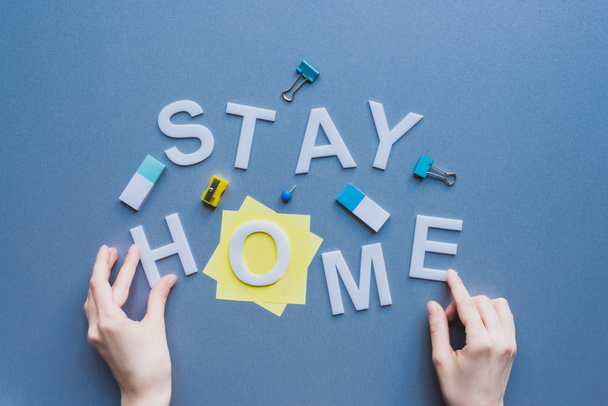 Top view of woman touching stay home lettering near erasers, binder clips and sticky notes on blue surface - Photo, Image