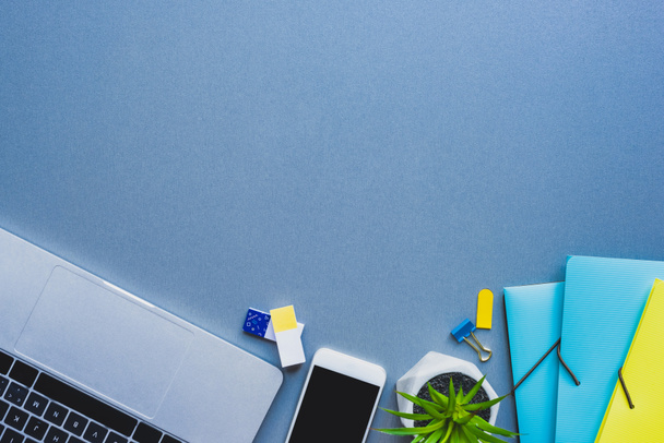 Top view of laptop, smartphone and plant near stationery on blue surface - Photo, Image