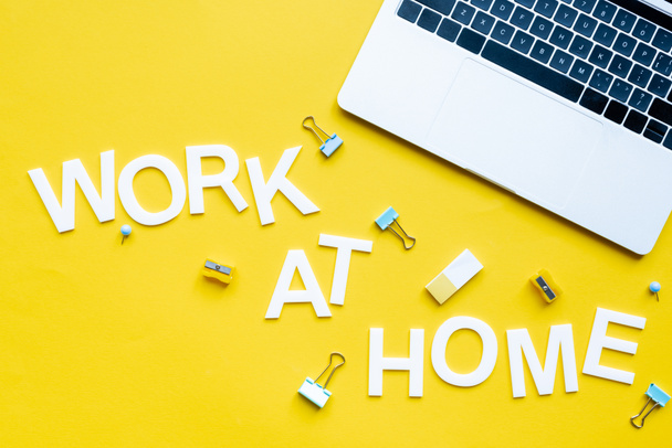 Vista superior do trabalho em casa lettering perto de laptop, grampos de aglutinante e apontadores de lápis na superfície amarela
 - Foto, Imagem