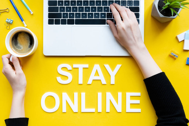 Bovenaanzicht van belettering verblijf online en vrouw met behulp van laptop en het houden van koffie op geel oppervlak - Foto, afbeelding