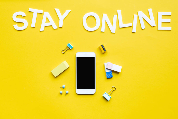 Bovenaanzicht van smartphone met blanco scherm in de buurt van briefpapier en blijf online belettering op geel oppervlak - Foto, afbeelding
