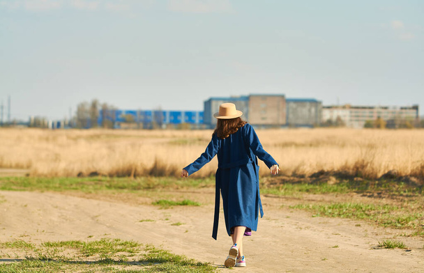 Szczęśliwa młoda dama w niebieskim płaszczu z beżowym kapeluszem biegnie na polu wczesną wiosną. - Zdjęcie, obraz
