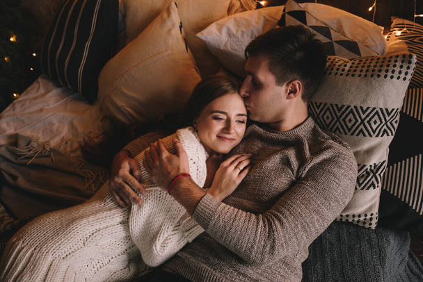 Couple on the bed in the bedroom. Dark interior. New Year's and Christmas. Hugs and kisses. Love. White sweater and high socks. Romantic meeting. A date lovers. Red thread on the wrist. - Фото, изображение