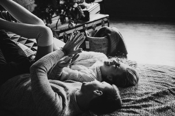 Black and white photo. Couple on the bed in the bedroom. Dark interior. New Year's and Christmas. Hugs and kisses. Love. White sweater and high socks. Romantic meeting. A date lovers. - Fotografie, Obrázek