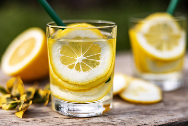 Летний напиток, стаканы воды, наполненные ломтиками лимонов, стоят на деревянной доске на газоне в солнечный теплый день, фон размыт, мелкая глубина резкости, выборочная фокусировка
. - Фото, изображение