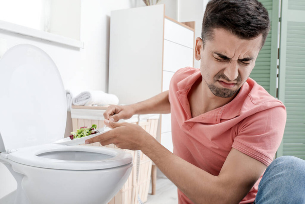 Displeased man suffering from anorexia near toilet bowl with salad on plate - Photo, Image