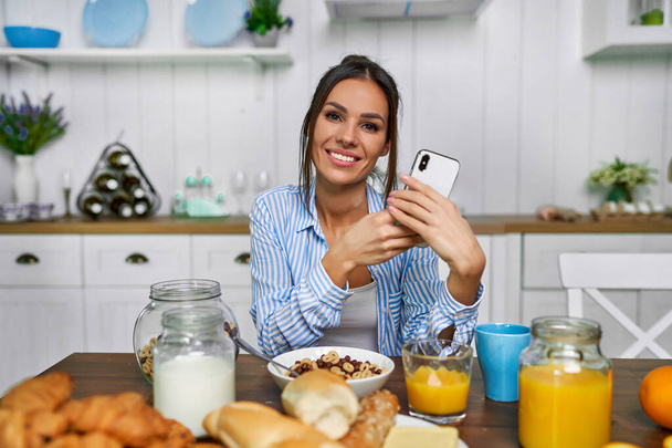 Beautiful attractive woman eating breakfast. Girl eating cereal with milk on the weekend and looking to the camera with phone - Foto, Imagem