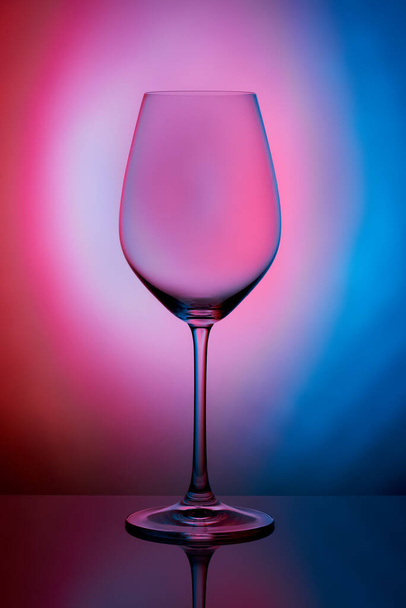 Кришталеве скло стоїть на відбиваючій поверхні на рожево-блакитному фоні. студійне фото. продукт. Красива тінь з келиха вина. макро фотографія
. - Фото, зображення