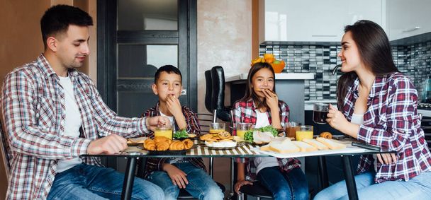 Veselí rodiče s dětmi při snídani doma, čtyři osoby sedí a jedí jídlo. - Fotografie, Obrázek