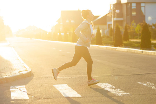 Νεαρή γυναίκα δρομέας, αθλητής τρέχει στο δρόμο της πόλης στο φως του ήλιου. Όμορφη καυκάσια γυναίκα εκπαίδευση, ακούγοντας μουσική - Φωτογραφία, εικόνα