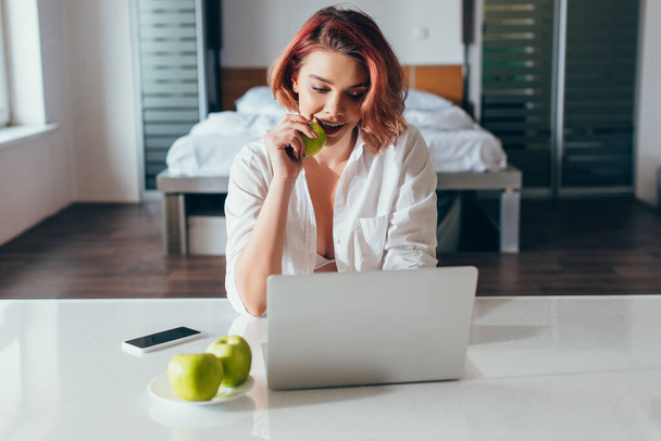 happy girl talking eating apple while using laptop at home on self isolation - Valokuva, kuva
