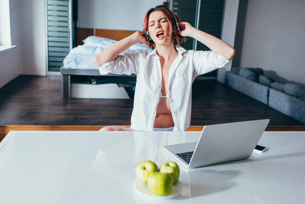 emotivní dívka poslouchá hudbu ve sluchátkách a zpívá doma s laptopem a jablky na karanténě - Fotografie, Obrázek