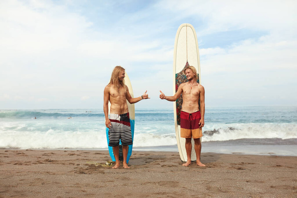 Surfeando. Jóvenes surfistas de pie en Sunny Beach. Hombres sonrientes mostrando el pulgar hacia arriba cerca de las tablas de surf en arena. Estilo de vida activo, Deporte acuático en hermoso fondo del océano
. - Foto, Imagen