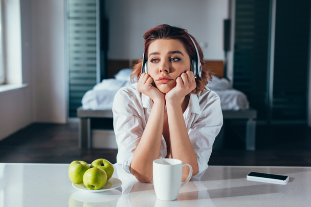 upset girl listening music with headphones on kitchen with coffee, apples and smartphone during self isolation - Fotó, kép