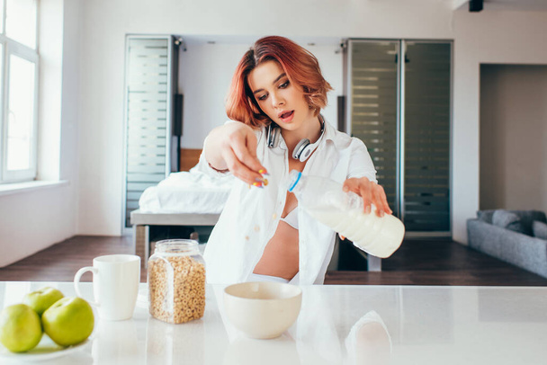 όμορφο κορίτσι με ακουστικά ρίχνει γάλα σε νιφάδες καλαμποκιού για πρωινό κατά τη διάρκεια της αυτο-απομόνωσης - Φωτογραφία, εικόνα