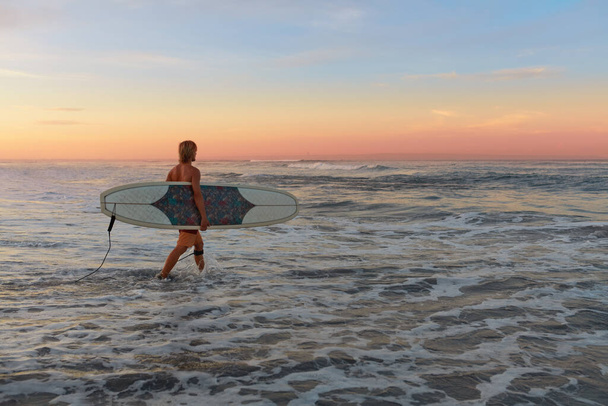 Surfer. Surfing Man With White Surfboard Walking In Ocean. Water Sport For Active Lifestyle. Beautiful Tropical Sunset At Sea. - Foto, afbeelding