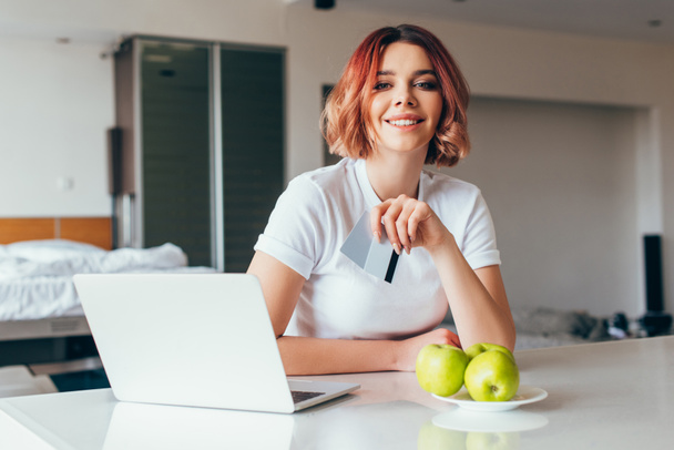 улыбающаяся девушка делает покупки онлайн с ноутбуком и кредитными картами на кухне с яблоками во время самоизоляции
  - Фото, изображение