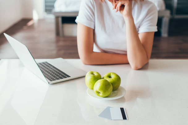 cropped view of girl shopping online with laptop and credit cards on kitchen with apples during quarantine  - Fotografie, Obrázek
