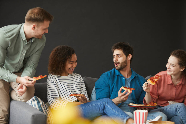 Grupo multiétnico de amigos comiendo pizza y aperitivos mientras disfrutan de una fiesta en casa sentados en un gran sofá sobre un fondo negro, copia el espacio arriba
 - Foto, Imagen
