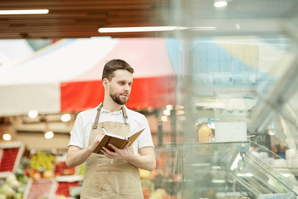 Retrato de la cintura hacia arriba del hombre barbudo que usa delantal y sostiene el cuaderno mientras hace el recuento de inventario en el supermercado, espacio de copia
 - Foto, imagen