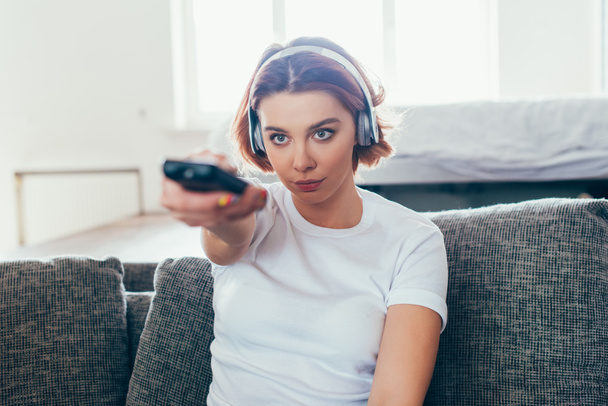atrakcyjna dziewczyna w słuchawkach trzymając pilota i oglądając telewizję w domu podczas samoizolacji - Zdjęcie, obraz