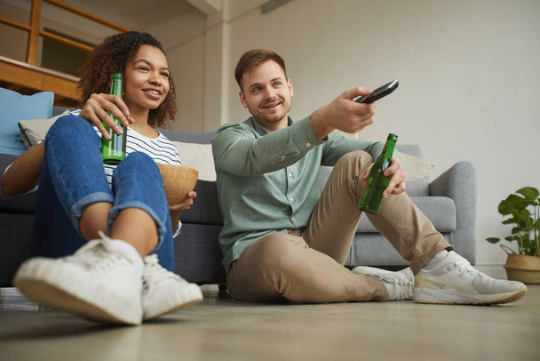 Plný portrét mladého smíšeného páru sledujícího doma televizi a popíjejícího pivo při sezení na podlaze v útulném bytě, kopírovací prostor - Fotografie, Obrázek