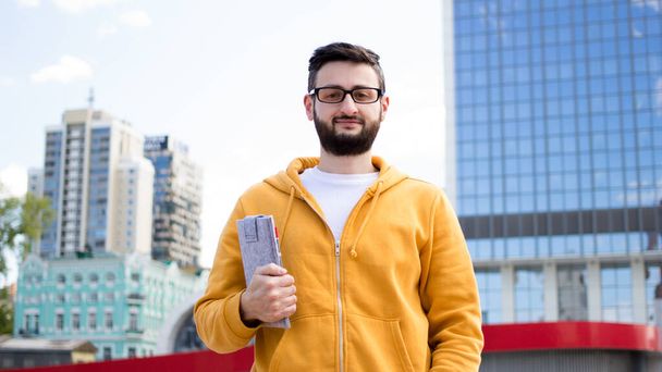 Młody, przystojny, brodaty mężczyzna w żółtej bluzie i okularach trzyma cyfrowy notatnik na szklanym tle budynku. Specjalista IT student hipster patrzy na kamery stojące na zewnątrz - Zdjęcie, obraz