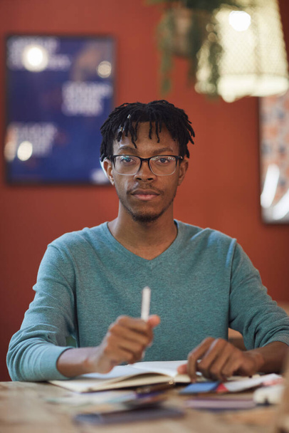 Retrato vertical del joven afroamericano mirando a la cámara mientras está sentado en la mesa contra la pared roja en la cafetería, espacio para copiar - Foto, Imagen