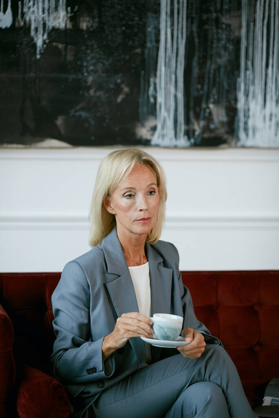 Retrato vertical de la moderna mujer de negocios madura sosteniendo la taza de café y mirando hacia otro lado pensativamente mientras se sienta en el sofá de terciopelo en la cafetería - Foto, Imagen