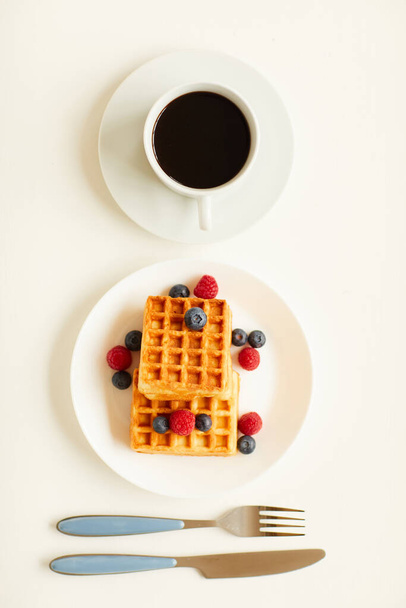 Top view at minimal composition of sweet dessert waffles with berry topping next to single cup of black coffee on white background, copy space - Фото, зображення