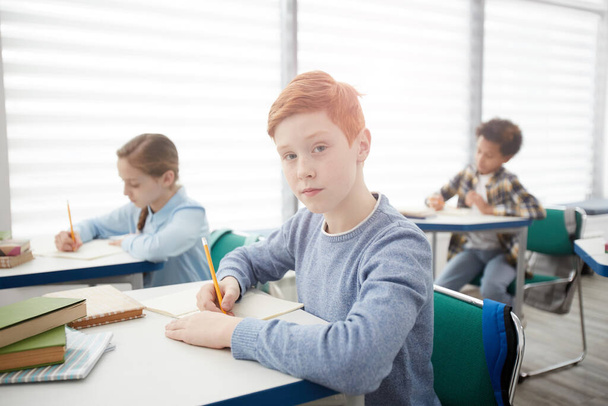 Πορτρέτο του σύγχρονου κοκκινομάλλη σχολιαρόπαιδο κάθεται στο γραφείο στην τάξη και κοιτάζοντας την κάμερα, ενώ κρατώντας σημειώσεις, αντίγραφο χώρο - Φωτογραφία, εικόνα
