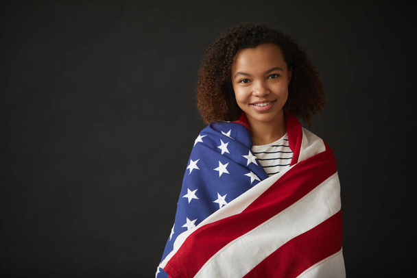 アフリカ系アメリカ人の少女がアメリカの国旗に包まれながら、黒い背景の中に立ってカメラで笑っている姿をウエストアップし、スペースをコピー - 写真・画像