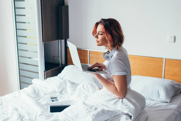 menina concentrada com fones de ouvido usando laptop na cama com smartphone e tablet digital no auto-isolamento
 - Foto, Imagem