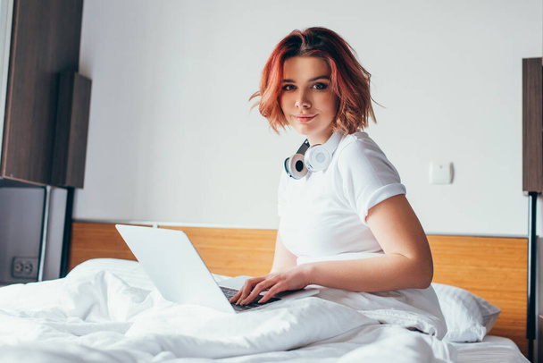 menina atraente com fones de ouvido usando laptop na cama em auto isolamento
  - Foto, Imagem
