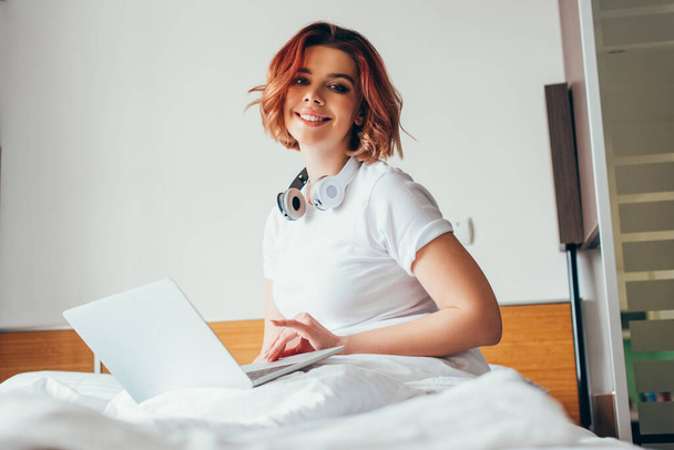 smiling girl with headphones using laptop in bed on self isolation - Photo, Image