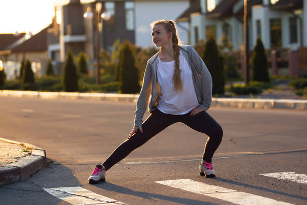 Young female runner, athlete is stretching before jogging in the city street in sunshine. Beautiful caucasian woman training, listening to music - Фото, изображение