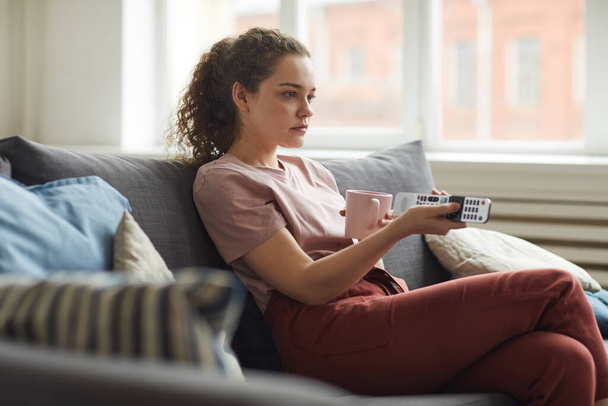 Widok z boku portret młodej kobiety oglądającej TV i trzymającej pilota podczas siedzenia na sofie w domu w przytulnym mieszkaniu, przestrzeń kopiowania - Zdjęcie, obraz