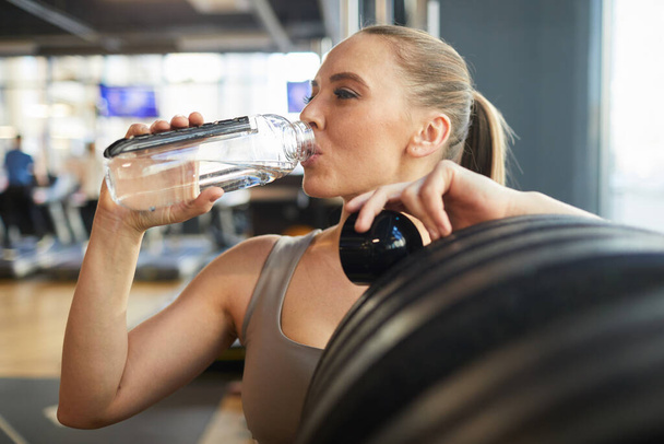 Retrato de cintura hacia arriba de una joven deportiva que bebe agua mientras se apoya en la máquina de ejercicios durante el entrenamiento de fuerza en el gimnasio moderno, espacio para copiar - Foto, Imagen