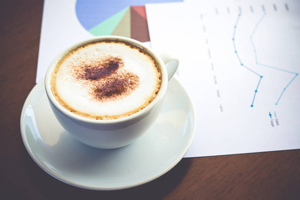 Eine weiße Kaffeetasse mit cremigem Schaum steht auf dem Schreibtisch mit Diagrammdaten. Büroarbeitskonzepte - Foto, Bild