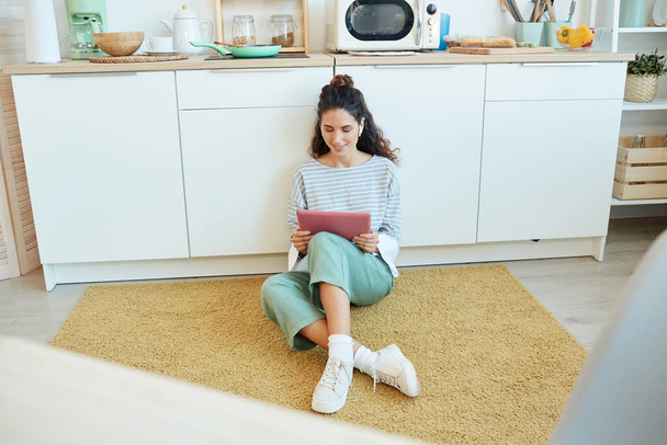 Young woman sitting on floor in kitchen room watching videos on her pink tablet computer - Foto, afbeelding