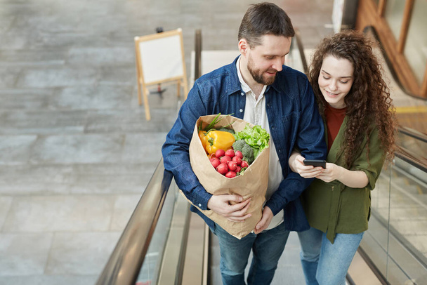 High angle portrait of happy modern couple holding bag with groceries while going up escalator in mall, copy space - Foto, Imagem