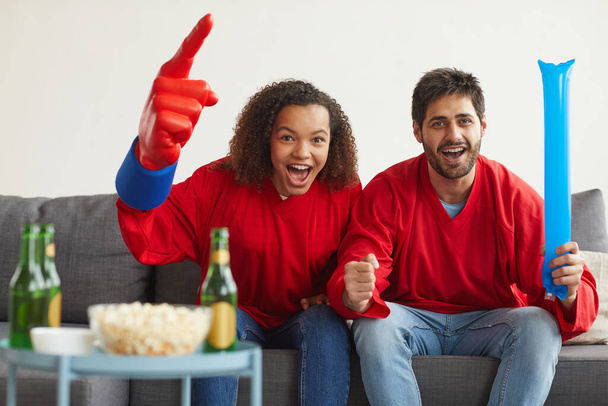 Πορτρέτο του σύγχρονου ζευγαριού μικτών αγώνων βλέποντας αθλητικό αγώνα στην τηλεόραση στο σπίτι και ζητωκραυγάζοντας συναισθηματικά φορώντας κόκκινες στολές της ομάδας, αντίγραφο χώρου - Φωτογραφία, εικόνα