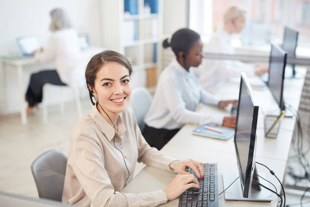 High angle portrait of female operator wearing headset and smiling at camera while sitting in row at call center, copy space - Photo, Image