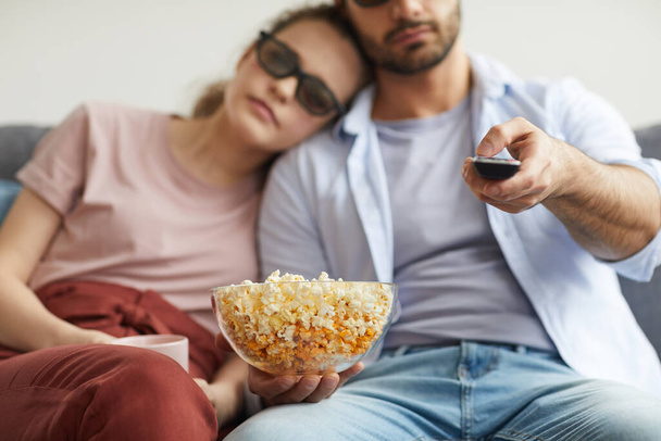 Blurred portrait of modern couple watching TV with stereo glasses while holding bowl of popcorn, focus on foreground, copy space - Fotografie, Obrázek