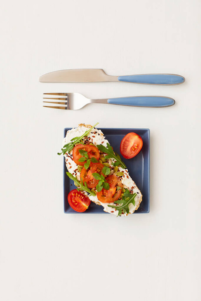 Top view at minimal composition of single bruschetta with cherry tomatoes and herbs next to knife and fork on white background, healthy breakfast and dieting concept, copy space - Photo, Image