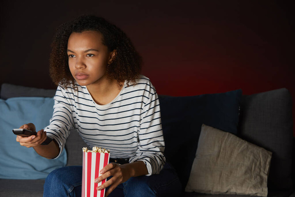 Portrait of young African American woman watching TV at home and holding remote control while sitting on couch in dark room - Photo, Image